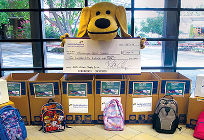 Sandy the Lab holding big check behind boxes filled with donated school supplies